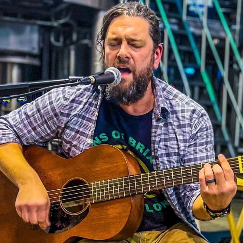 Man with a beard and closed eyes passionately plays an acoustic guitar and sings into a microphone. He wears a plaid shirt and sits on a stage.