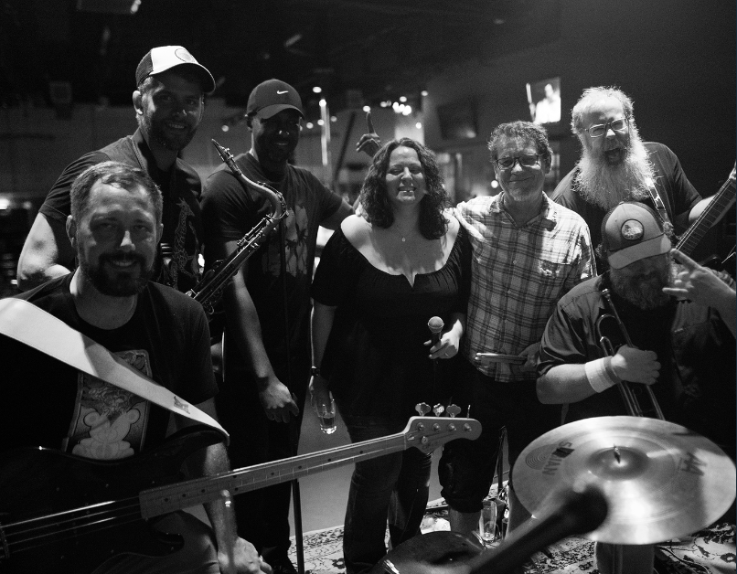 A diverse group of seven musicians poses on a dimly lit stage, holding instruments and smiling. The scene conveys creativity and camaraderie.